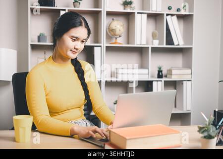 Young Asian woman using laptop at home office desk with books and coffee, studying online or working remotely. Smiling student in modern workspace, e- Stock Photo