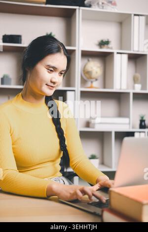 Young Asian woman using laptop at home office desk with books and coffee, studying online or working remotely. Smiling student in modern workspace, e- Stock Photo