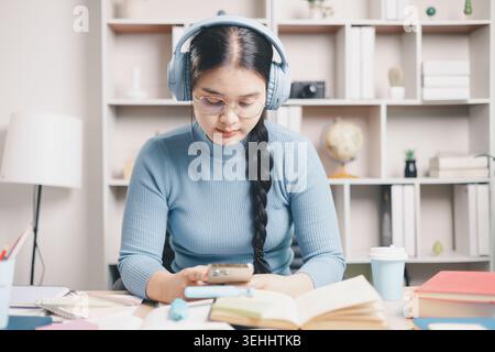 Young Asian woman in glasses using smartphone while studying with books and headphones at home desk. Mobile learning, online research, digital educati Stock Photo