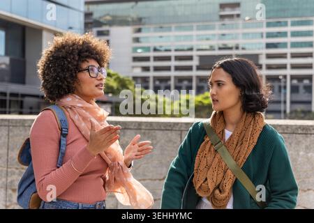 Diverse female friends standing and talking in urban plaza by concrete barrier with polka-dot scarf Stock Photo