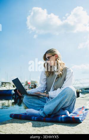 A woman sits on the ground near the water with a laptop. She is wearing casual clothes and looking at the screen. Boats are visible in the background Stock Photo
