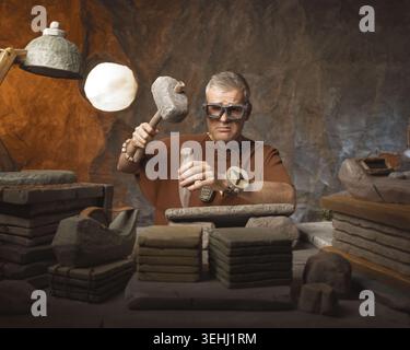 Frustrated office worker caveman sitting at his desk and hand carving inscriptions on stone tablets using a hammer and a chisel Stock Photo