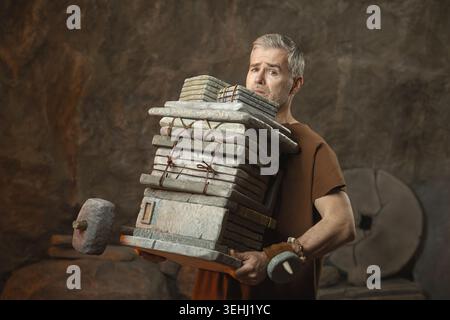 Funny portrait of a tired caveman office worker struggling to carry a huge stack of heavy stone slabs and folders. Humorous metaphor for being overwor Stock Photo