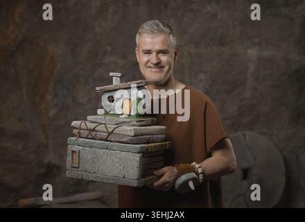 Happy caveman holding a stack of stone documents with a miniature stone house model on top. Humorous concept of a prehistoric architect or real estate Stock Photo