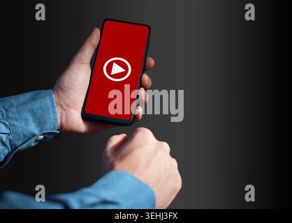 Person holding a smartphone, the screen displaying a red background with a white play button, indicating video content or streaming Stock Photo
