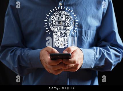 Man holding smartphone in hands, a glowing lightbulb composed of various business and technology icons hovering above. Technology idea creation and de Stock Photo