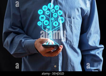 Businessman holding a smartphone, a digital tree of internet of things icons emerging, representing connectivity, networking, and data management Stock Photo