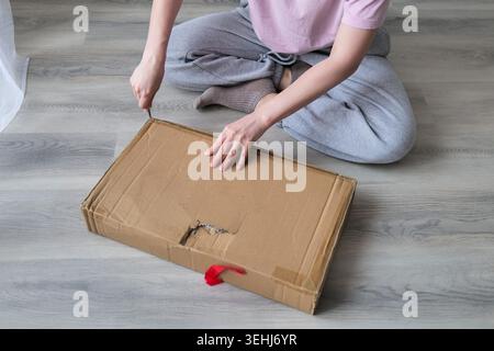 Woman Opening Cardboard Box with Knife While Sitting on Floor at Home Stock Photo