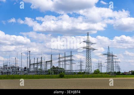 View of Kraftwerk Irsching power plant substation and high-voltage transmission towers standing in a field under a blue sky with clouds in Vohburg an Stock Photo
