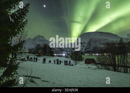 View of the vibrant Aurora Borealis dancing over snow-covered mountains and people gathered in the village of Lakselvbukt, Troms, Norway. Stock Photo