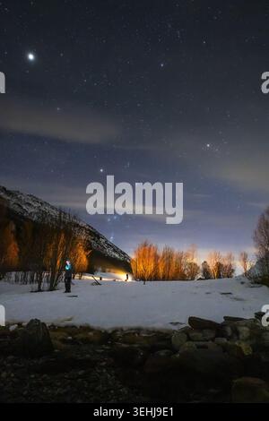 View of a person standing in a snowy field under a starry night sky with illuminated trees and a mountain ridge in Lakselvbukt, Troms, Norway. Stock Photo