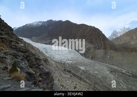 View of a massive white glacier flowing between rugged mountain peaks under a cloudy sky Gilgit-Baltistan, Pakistan. Stock Photo