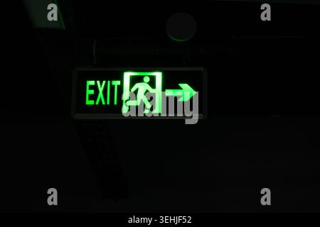 Illuminated green exit sign with a running man pictogram and direction arrow hanging in a dark corridor or public building. Safety, evacuation, and em Stock Photo
