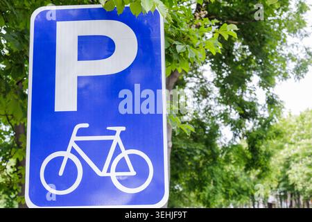 A blue bicycle parking lot icon sign located on the left side of the frame under a green tree canopy. Right side features soft blurred bokeh backgroun Stock Photo