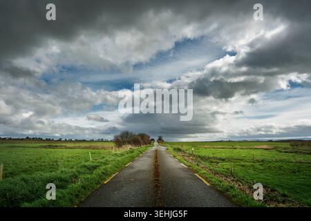 Narrow country road leading through green fields under a dramatic cloudy sky in County Kerry, Ireland. Stock Photo