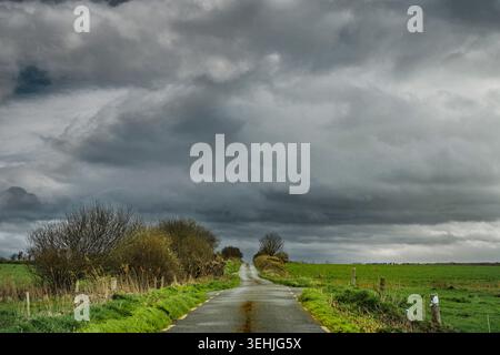 Dramatic rural landscape of a narrow Irish boreen road leading toward heavy rain clouds and stormy weather. Stock Photo