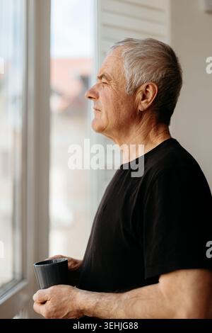 Senior man gazing thoughtfully through window while holding coffee cup Stock Photo