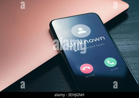 Spam call warning on smartphone screen with unknown caller and suspected scam alert beside laptop. Mobile fraud, robocall protection and cybersecurity concept. High quality photo Stock Photo