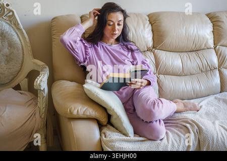 Nice woman reading book on sofa at home. Mature brunette wearing domestic velour pants and long sleeve sweatshirt Stock Photo