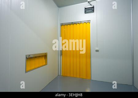 Empty industrial cold room interior featuring white sterile wall grey floor bright yellow plastic strip curtain door and small window conveying quiet Stock Photo
