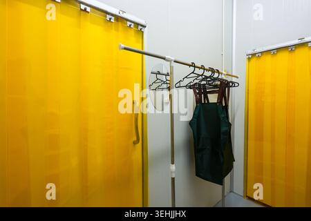 Empty room features bright yellow plastic strip curtain and metal clothing rack holding dark green apron on black hanger portraying calm organized ste Stock Photo