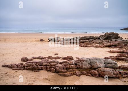 Natural pink sandstone sculptures dot the golden sand of a Breton beach. The calm ocean blends with a gray sky on the horizon, creating an atmosphere Stock Photo