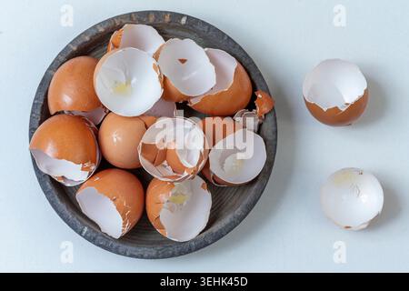 Brown eggshells are piled inside a dark grey bowl, with two additional shells resting on the white surface beside it. Stock Photo