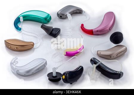 Various models and colors of hearing aids, including behind-the-ear and in-ear devices, arranged on a white surface. Stock Photo