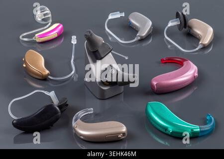 Various models and colors of hearing aids, including behind-the-ear and in-the-ear devices, arranged on a reflective grey surface. Stock Photo