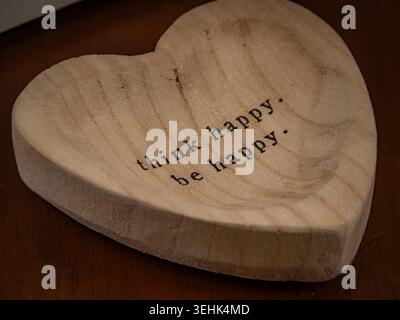 Close up of a heart shaped light wood tray engraved with the inspirational text think happy be happy resting on a dark wooden surface Stock Photo