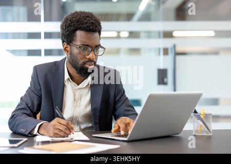 Young man wearing business suit and glasses, working diligently at a modern office desk, typing on his laptop and simultaneously writing down important notes in a professional workspace Stock Photo