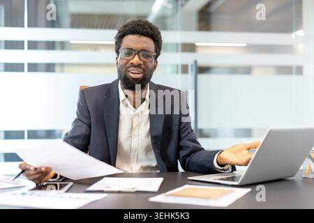 Professional man showing confusion and frustration, holding documents and gesturing towards a laptop while experiencing work stress or a challenging problem in a modern office setting Stock Photo