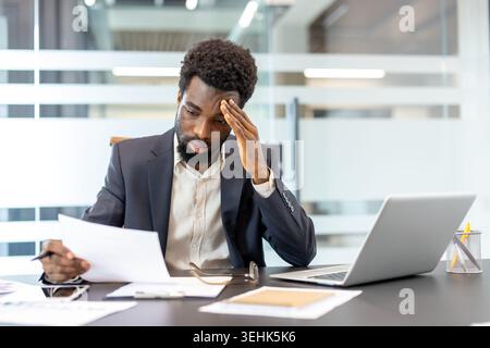 Focused african american businessman at his desk, reviewing paperwork with a concerned, tired expression while managing finance documents and deadline pressure in a corporate office Stock Photo