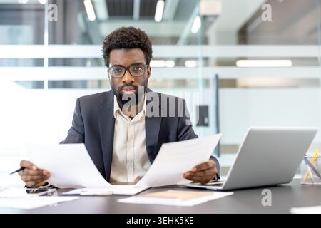 Young businessman analyzing financial reports and paperwork while sitting at his desk with a laptop, focusing on important data for strategic decision-making and planning Stock Photo