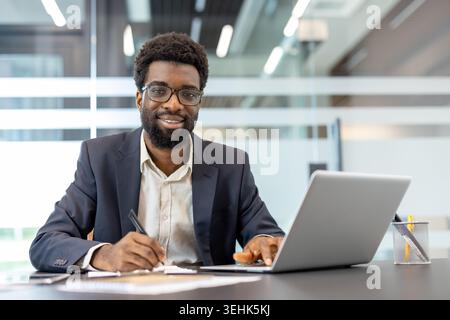 Young businessman in suit smiles confidently at camera while working at a modern office desk, taking notes on documents beside his laptop, conveying leadership and productivity Stock Photo