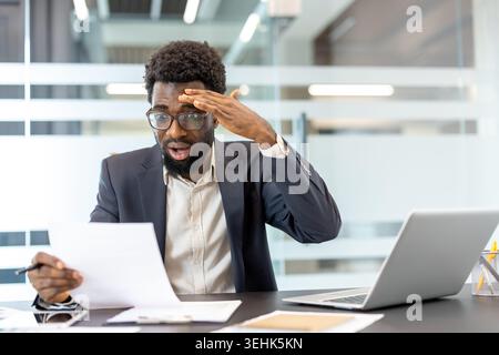 Young businessman experiencing shock and disappointment while reading financial documents, holding his head in disbelief after discovering a mistake or problem with the paperwork in a modern office Stock Photo