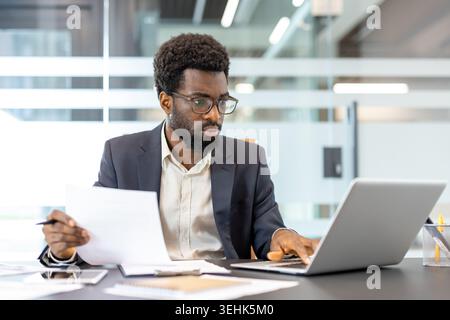 Professional businessman with eyeglasses checking financial documents and typing on a laptop, actively working and focusing on tasks at a modern office desk Stock Photo