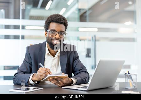 African american businessman working at his desk, smiling at the camera, and writing notes in a notebook while using a laptop and tablet in a modern office environment Stock Photo