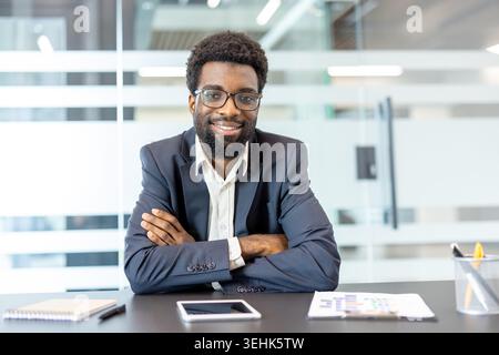 Smiling multiracial business man wearing glasses and a suit, sitting at his office desk with arms crossed, conveying confidence and professionalism in a contemporary workspace Stock Photo