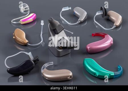 Various models and colors of hearing aids, including behind-the-ear and in-the-ear devices, arranged on a reflective grey surface. Hearing aids. Bsip 016938 147 Stock Photo
