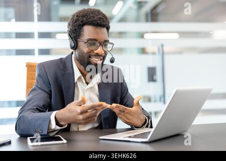 Confident young business professional wearing a headset and glasses. Actively engaging in a video conference from an actual office. Communicating with clients or colleagues while gesturing with hands Stock Photo