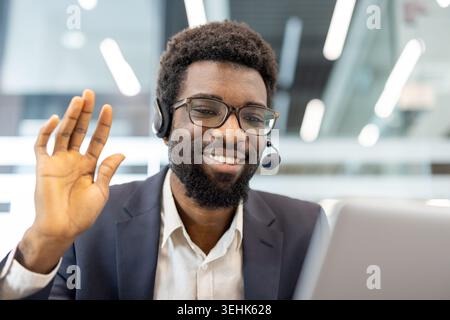 Smiling man wearing glasses and a headset, raising his hand in a wave while participating in a virtual meeting or online support session from an office environment, engaging in customer service Stock Photo