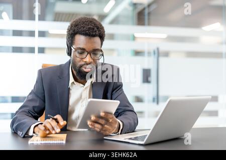 Professional man wearing a headset and spectacles, actively engaging with a digital tablet while taking notes, providing customer service and support in a modern office environment Stock Photo