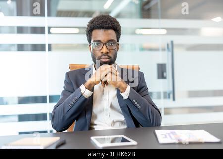 Confident businessman with serious expression sitting at a conference table, hands clasped together, pondering solutions or making strategic decisions in a contemporary office environment Stock Photo