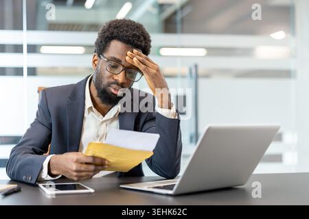 Stressed businessman in a modern office receiving bad news in a letter, experiencing worry, debt, and loss while looking at a laptop and smartphone on his desk Stock Photo