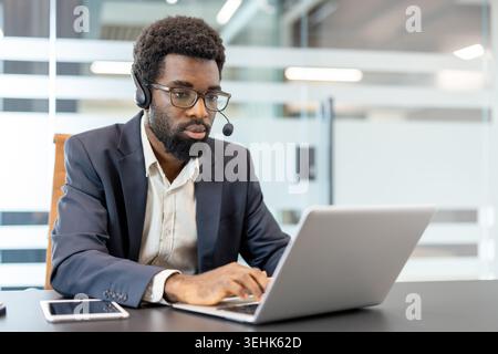 Young male employee providing customer service assistance, actively typing on a laptop with a headset, demonstrating dedicated support and communication in a modern workspace Stock Photo