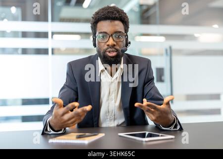 Professional man wearing a headset and glasses, engaging in a virtual video conference, communicating effectively while gesturing with hands in a modern office environment Stock Photo