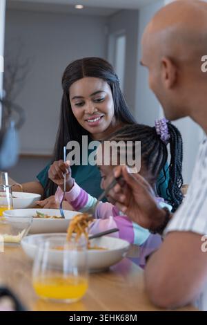 African American family eating at home table, mom in teal smiling while daughter eating pasta Stock Photo