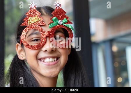 Indian female child smiling and wearing red glitter Christmas-tree glasses at cafe windows Stock Photo