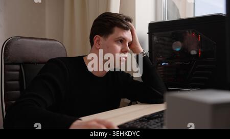 A young, overworked freelancer feeling tired and stressed at his desk. He yawns from exhaustion and lack of sleep, showing signs of burnout while work Stock Photo